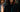 Photorealistic editorial portrait of Rebel Mythic standing in a rain-soaked downtown street at night
